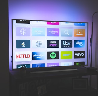 A flat-screen television is displayed in a dimly lit room, showcasing a variety of streaming service apps on its screen, such as Netflix, Prime Video, and Spotify. The TV is positioned on a black stand beside a tall floor lamp, casting a soft ambient light on the surrounding area.
