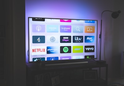 A flat-screen television is displayed in a dimly lit room, showcasing a variety of streaming service apps on its screen, such as Netflix, Prime Video, and Spotify. The TV is positioned on a black stand beside a tall floor lamp, casting a soft ambient light on the surrounding area.