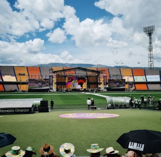 A sports stadium with a large stage set up on the field, hosting an outdoor event. Spectators wearing colorful hats sit in the foreground. The seating areas display vibrant colors, and a clear blue sky with scattered clouds is visible. Large screens display graphics related to the event.