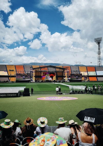 A sports stadium with a large stage set up on the field, hosting an outdoor event. Spectators wearing colorful hats sit in the foreground. The seating areas display vibrant colors, and a clear blue sky with scattered clouds is visible. Large screens display graphics related to the event.