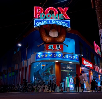 A vibrant, neon-lit facade of a gaming and sports establishment at night, featuring a large glove holding a baseball above the entrance. The building is adorned with colorful signs in both English and Japanese, with various advertisements and an illuminated sign that says 'ROX DOME'. Several people are walking towards the entrance, and bicycles are parked along the side.