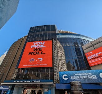 A cityscape image featuring the entrance of Madison Square Garden with large digital advertisements on the building facade. The advertisements promote a partnership with the New York Knicks and DoorDash, featuring bold typography and vibrant orange backgrounds. The iconic Empire State Building is visible in the background, and the clear blue sky adds to the urban setting. The signage for Pennsylvania Station is prominently displayed below.