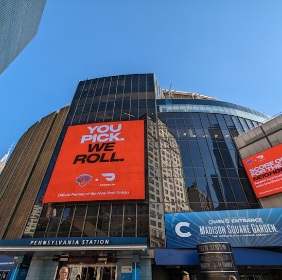 A cityscape image featuring the entrance of Madison Square Garden with large digital advertisements on the building facade. The advertisements promote a partnership with the New York Knicks and DoorDash, featuring bold typography and vibrant orange backgrounds. The iconic Empire State Building is visible in the background, and the clear blue sky adds to the urban setting. The signage for Pennsylvania Station is prominently displayed below.