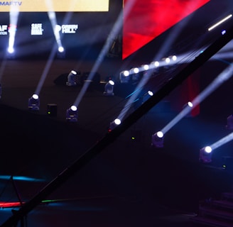 An array of bright stage lights beams across a dark arena, creating a dynamic atmosphere with a large screen displaying various logos and text. A camera crane is visible in the foreground, suggesting a professional event setup.