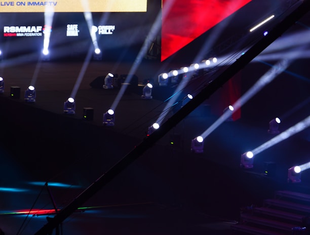 An array of bright stage lights beams across a dark arena, creating a dynamic atmosphere with a large screen displaying various logos and text. A camera crane is visible in the foreground, suggesting a professional event setup.