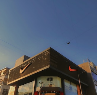 A modern building with predominantly gray and black tones featuring a large, well-known sports logo in bright orange. The facade includes glass panels displaying large advertisements. The sky is clear and blue, suggesting a sunny day. Nearby utility poles and wires are also visible.