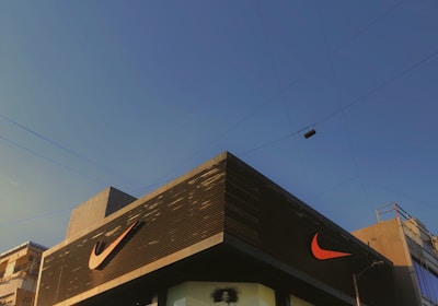 A modern building with predominantly gray and black tones featuring a large, well-known sports logo in bright orange. The facade includes glass panels displaying large advertisements. The sky is clear and blue, suggesting a sunny day. Nearby utility poles and wires are also visible.